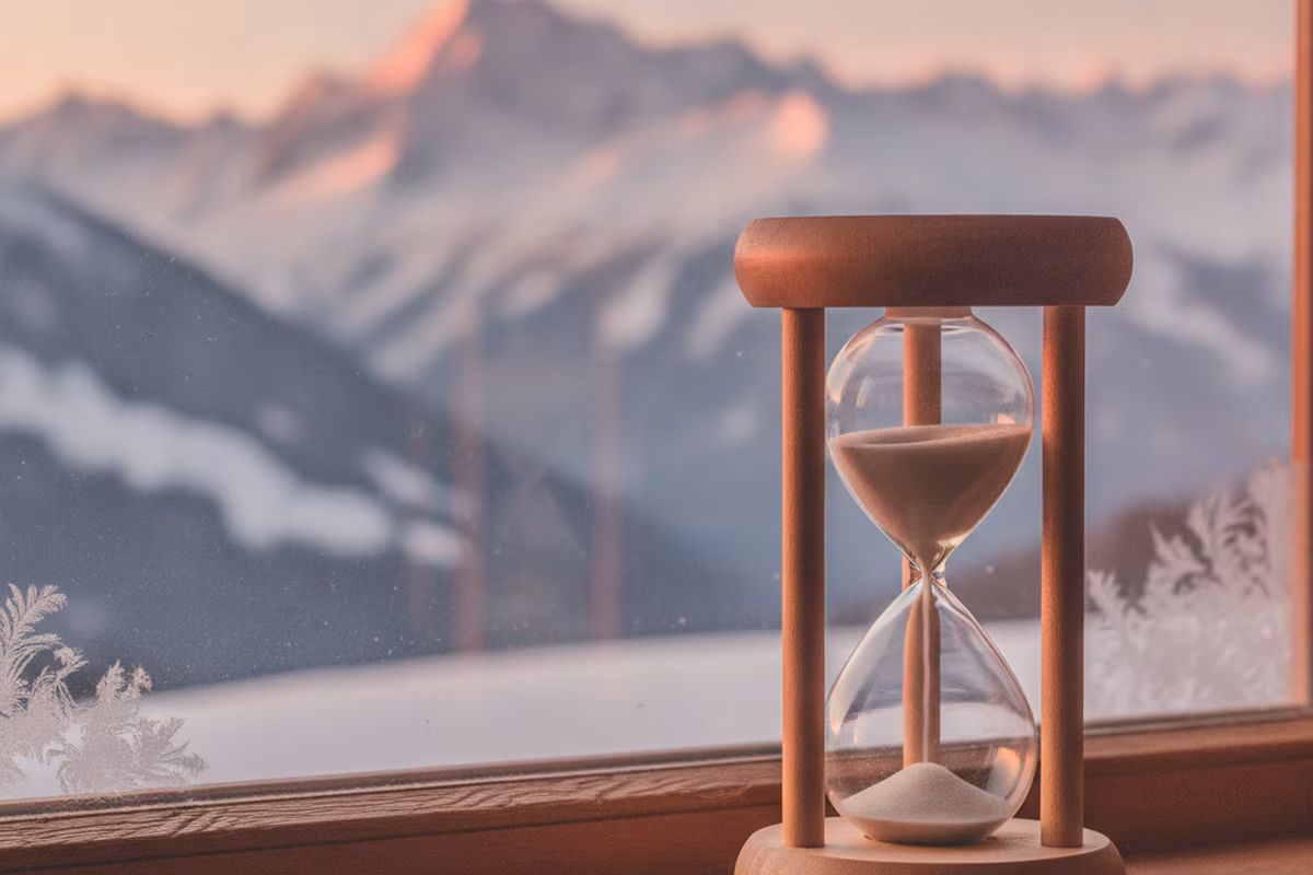 Hourglass standing on a sunlit wooden chalet windowsill with mountains beyond